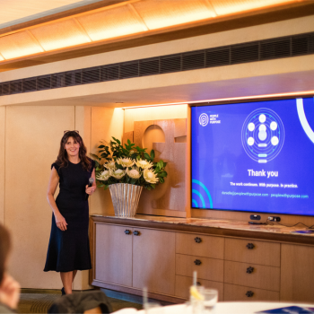 Woman standing beside a floral arrangement, presenting to a screen displaying a "Thank you" slide with a circular logo graphic.